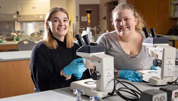 Students in lab setting sitting behind microscopes, looking at camera smiling