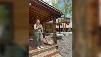 Person standing with a broom in front of the Children's Building, surrounded by trees and picnic tables.