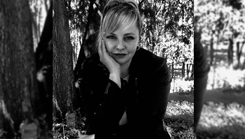 Person with short hair, leaning on their hand, smiling gently while sitting in a park.