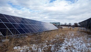 Rows of solar panels in a field with light snow cover under a cloudy sky.