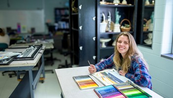Person smiling at a table with colorful pastel chalks in a classroom.