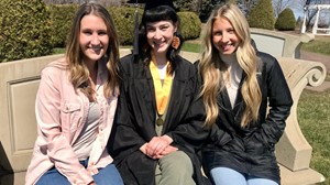 Three individuals seated on a bench outdoors, smiling, with one in graduation attire. They are in a park-like setting with a structure resembling a bridge in the background.