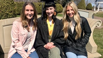 Three individuals seated on a bench outdoors, smiling, with one in graduation attire. They are in a park-like setting with a structure resembling a bridge in the background.