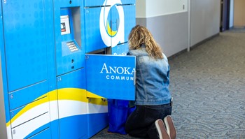 A person is crouched in front of a vibrant blue locker system labeled 'Anoka Community', seemingly interacting with it or retrieving items.