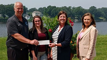 A group of four people standing outdoors. One person is handing a check to another, while two others stand beside them. 