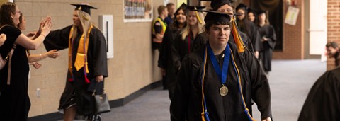 A group of graduates in black caps and gowns walk down an indoor hallway during a ceremony.